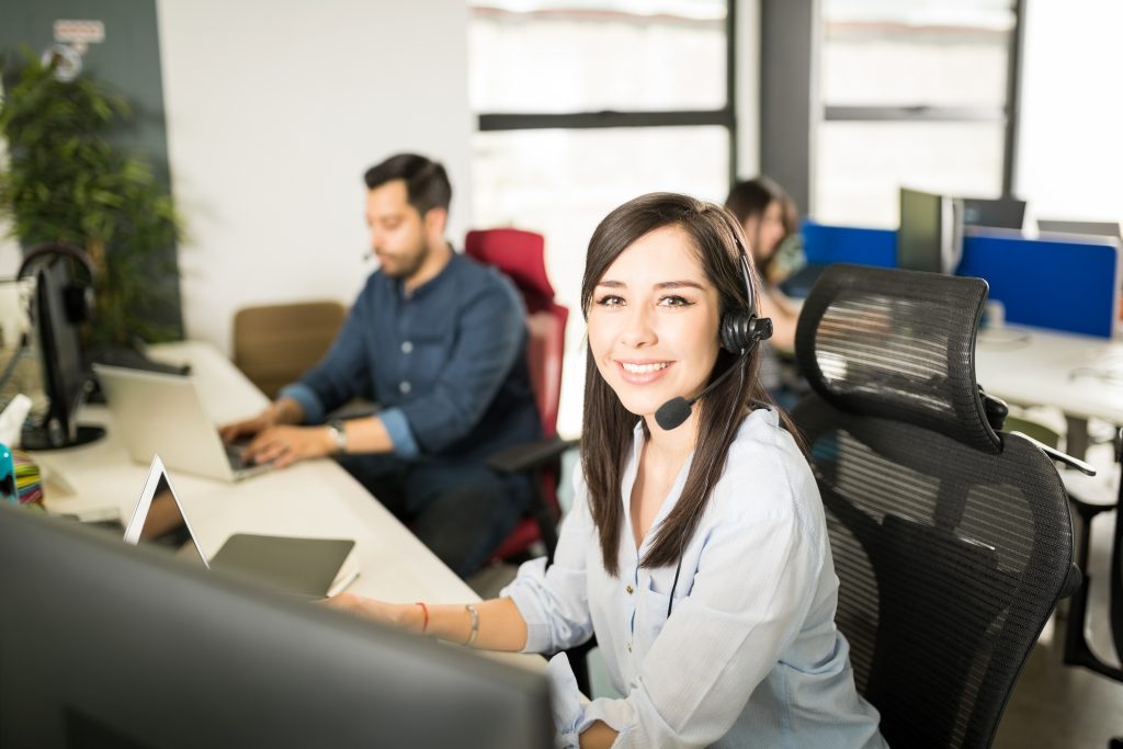 Portrait of pretty young Latin woman wearing a headset sitting at her office desk with coworkers in background